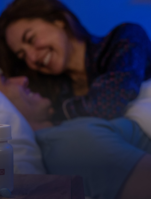 A couple is smiling in bed with Rex MD Viagra medication in front of them on a night stand